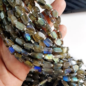 Pode incluir: Fios de contas retangulares de labradorite em tons de cinza, marrom e azul. As contas são enfiadas em um fio branco, criando um efeito em camadas. As pedras preciosas polidas são seguradas em uma mão.
