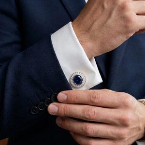 Könnte beinhalten: Ein runder, silberfarbener Manschettenknopf mit einem tiefblauen, kreisförmigen Mittelstein, der von einem Kranz aus kleinen, klaren Steinen umgeben ist, befestigt an der weißen Manschette eines dunkelblauen Sakkos mit vier schwarzen Knöpfen.