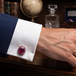 May include: A silver-toned oval cufflink featuring a large central red gemstone surrounded by a halo of smaller red stones, secured on a crisp white French cuff of a navy blue blazer sleeve, showcasing a classic and refined accessory design.