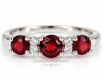 May include: A silver-toned ring featuring three round-cut red gemstones and four smaller clear round-cut stones set between them. The text LuxuryGemsJewelry is printed below the ring with a diamond-shaped icon and horizontal lines.