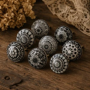 May include: Nine black and white ceramic cabinet knobs with detailed patterns. The knobs are circular with brass stems, displayed on a wooden surface. The designs feature floral and geometric patterns. The knobs are suitable for furniture.
