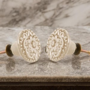 May include: Two cream-coloured ceramic cabinet knobs with floral patterns. Each knob features a brass screw and a dark green base. The knobs are shown on a grey surface with a marble backdrop.