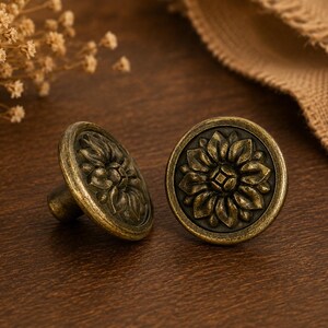 May include: Two antique brass cabinet knobs with a floral design. The knobs are round with a raised floral pattern in the centre and a dark, aged finish. The knobs are set against a dark wood surface.