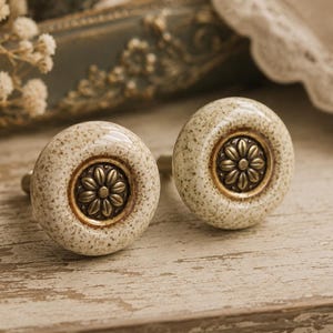 May include: Two ceramic cabinet knobs with a speckled beige finish and a brass flower design in the center. The knobs are set against a rustic, light-colored wooden surface, suggesting a vintage or farmhouse style.