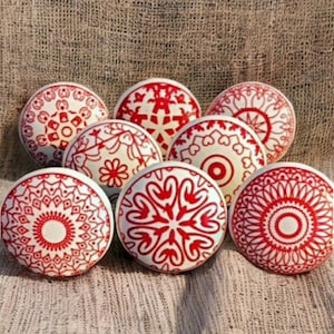 Op de afbeelding: Set van acht witte keramische knoppen met rode geometrische ontwerpen. De knoppen hebben een verscheidenheid aan patronen, waaronder sneeuwvlokken, harten en cirkels.
