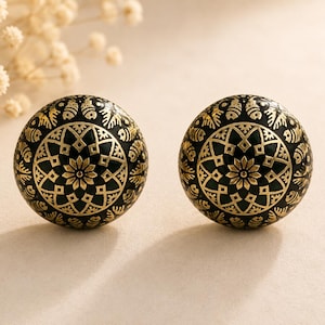 May include: Two black and gold decorative spheres with detailed floral and geometric designs. The spheres feature a dark base with gold detailing, possibly used as ornaments or decorative items. They are presented on a neutral surface, with a soft focus background.
