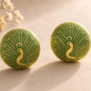 May include: Two circular cabinet knobs featuring a peacock design. The knobs are gold with a green peacock illustration. The peacock's tail feathers are fanned out, and the bird is facing sideways. The knobs are set against a neutral backdrop.