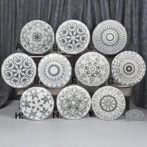 Może przedstawiać: Kolekcja jedenastu białych ceramicznych gałek do szafek z różnymi szarymi geometrycznymi wzorami. Gałki są okrągłe i mają wzory takie jak kwiaty, gwiazdy i symetryczne kształty. Gałki są wystawione na drewnianej powierzchni.