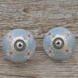 Op de afbeelding: Twee lichtblauwe keramische kastknoppen met een zilveren metalen basis en witte en oranje bloemmotieven.