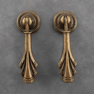 May include: A pair of antique bronze drawer pulls. Each pull has a circular top with a detailed design and an elongated, decorative handle. These metal pulls are designed for use on furniture.