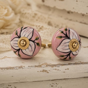 Può includere: Due pomelli rotondi in ceramica rosa con un motivo floreale. I pomelli presentano un fiore bianco con contorni neri e ferramenta dorata. Lo sfondo è una superficie bianca invecchiata.