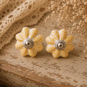 May include: Two decorative flower-shaped cabinet knobs. The knobs are white with yellow and orange speckled patterns. Each knob has a silver-coloured metal centre. The knobs are on a distressed wooden surface.
