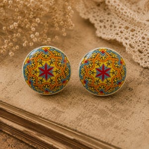 May include: Two round cabinet knobs with a colorful geometric design. The knobs feature a yellow background with a red, blue, and orange starburst pattern. The knobs are likely made of ceramic or porcelain.