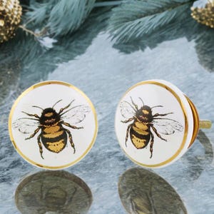 May include: Two white ceramic cabinet knobs with a gold rim and a detailed bee illustration. The bee is black and yellow. The knobs have a gold screw and are suitable for furniture or decor.