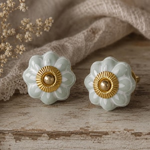 Könnte beinhalten: Zwei hellgrüne florale Schrankknöpfe mit goldenen Akzenten. Die Knöpfe haben ein blütenartiges Design mit einem goldenen Zentrum und einem gewellten Rand. Sie sind vor einem neutralen Hintergrund platziert.