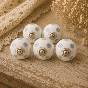 May include: Five white ceramic cabinet knobs with a silver base and light blue polka dots. The knobs are arranged on a wooden surface, with a lace trim and dried flowers in the background. These decorative knobs are ideal for furniture.