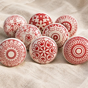 Puede incluir: Ocho pomos de cerámica blanca con patrones decorativos rojos. Los diseños incluyen motivos florales, geométricos y mandala. Los pomos se muestran sobre una tela de color neutro. Cada pomo mide aproximadamente 4 cm de diámetro.