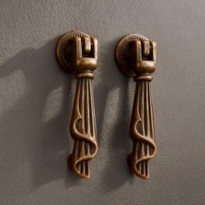 Può includere: Due maniglie antiche in bronzo per mobili. Ogni maniglia ha un'impugnatura decorativa e scanalata con un design stilizzato. Le maniglie sono montate su una superficie grigia. Le maniglie sono probabilmente per un armadio o un cassetto.