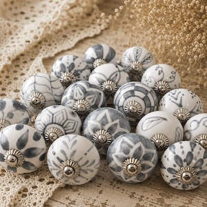 Può includere: Una collezione di pomelli decorativi in ceramica per mobili. I pomelli sono bianchi con motivi floreali e geometrici grigi. Ogni pomello ha una base in metallo color argento. I pomelli sono disposti su una superficie di pizzo beige.