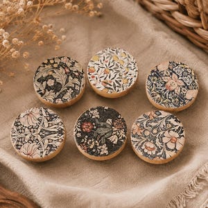 Puede incluir: Seis posavasos redondos de madera con estampados florales en varios colores, incluyendo crema, negro y rojo. Los posavasos están dispuestos sobre una superficie de lino beige, con flores secas y una cesta de mimbre en el fondo. Diámetro: aprox. 9 cm.