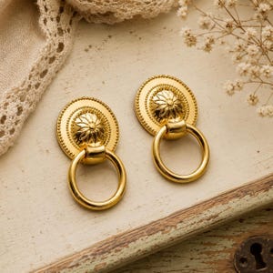 Könnte beinhalten: Zwei goldfarbene Schrankgriffe mit floralem Design. Jeder Griff hat eine kreisförmige Rückplatte mit erhabenem Blumenzentrum und einem Ringgriff. Die Griffe werden auf einer hellen, abgenutzten Holzoberfläche präsentiert.