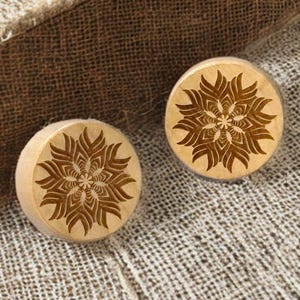 Op de afbeelding: Twee ronde houten pluggen met een gedetailleerd bloemmotief in het oppervlak gesneden. De pluggen zijn lichtbruin en liggen op een textiel met neutrale tint. Het bloemmotief is donkerder bruin.