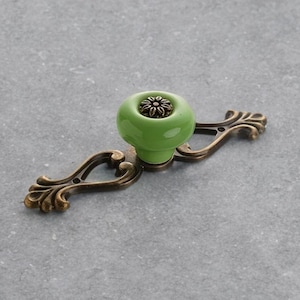 Op de afbeelding: Een decoratieve kastgreep met een heldergroene keramische knop en een bronzen metalen achterplaat. De knop heeft een bloemmotief aan de bovenkant. De achterplaat heeft een sierlijk, gebogen ontwerp.