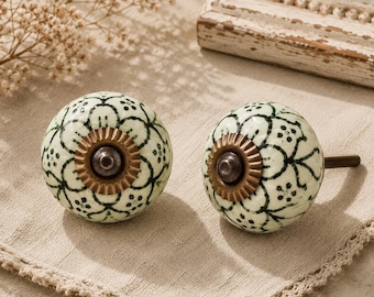 May include: Two decorative ceramic cabinet knobs. Each knob is round, with a cream-colored base and a black floral pattern. They have a bronze-colored metal base and a screw for installation. The knobs are designed for furniture and home decor.