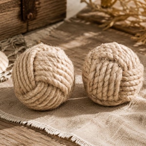 May include: Two decorative rope balls, each meticulously crafted with thick, natural-colored rope. The balls are knotted in a round shape, resting on a beige linen cloth. A seashell and wooden chest are in the background, suggesting a nautical theme.