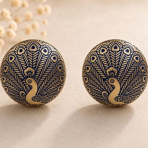 May include: A pair of gold-coloured, round earrings featuring a navy blue peacock feather design. Each earring has a detailed pattern and a curved peacock shape. The background is a neutral beige colour.