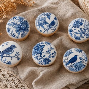 Op de afbeelding: Zes ronde kastknoppen met witte en blauwe bloemen- en vogelontwerpen. De knoppen hebben een houten basis en een keramische bovenkant. De ontwerpen omvatten verschillende vogels en bloempatronen in een klassiek blauw-wit kleurenschema.
