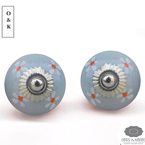 Puede incluir: Dos pomos de armario de cerámica azul claro con una base de metal plateado y diseños de margaritas blancas y naranjas.