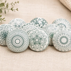 Puede incluir: Un conjunto de ocho pomos de cerámica para armarios con intrincados diseños florales y geométricos en color verde azulado y blanco. Los pomos están dispuestos sobre una superficie de lino neutro, mostrando sus diseños decorativos.