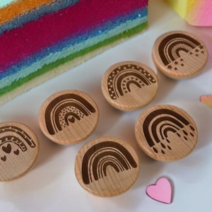 Peut inclure: Boutons décoratifs en bois sur le thème de l'arc-en-ciel, avec divers motifs gravés, notamment des cœurs et des étoiles. Les boutons sont disposés sur une surface blanche, avec un objet coloré en arrière-plan. Des confettis en forme de cœur roses et pêche sont éparpillés.