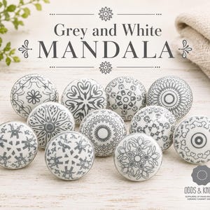 May include: A selection of grey and white mandala ceramic cabinet knobs. Each knob has a unique design, including floral and geometric patterns. The knobs are hand-painted and displayed on a light wooden surface. The text "Grey and White MANDALA" is at the top.