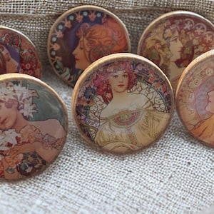 Può includere: Una collezione di sottobicchieri rotondi in legno con illustrazioni in stile Art Nouveau di donne con elementi floreali e decorativi. I sottobicchieri hanno un bordo in legno chiaro e sono disposti su una superficie strutturata.