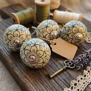 May include: Five cream-colored ceramic cabinet knobs with a green floral design and silver hardware. A vintage key, blank tag, and spools of thread are arranged on a wooden surface, suggesting a craft or home decor theme.