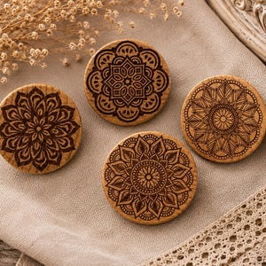 Könnte beinhalten: Vier runde Untersetzer aus Holz mit aufwendigen Mandala-Designs, die in ihre Oberflächen geätzt sind. Die Untersetzer sind hellbraun, die Designs sind dunkelbraun. Sie sind auf einem beigen Tuch angeordnet, mit getrockneten Blumen im Hintergrund.