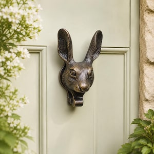 May include: A bronze-colored door knocker shaped like a rabbit's head. The knocker is mounted on a light green door with a vertical panel design. The rabbit has detailed fur texture and a curved handle below its face.