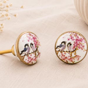 May include: Two decorative cabinet knobs featuring a bird and floral design. The knobs are white with a gold rim and stem. The design shows two birds perched on a branch with pink flowers.