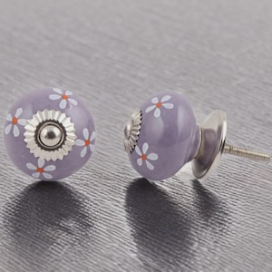 Puede incluir: Dos pomos de cerámica lavanda con diseños de margaritas blancas y centros naranjas. Cada pomo tiene una base y un tornillo de metal plateado. Pomos decorativos para muebles o decoración del hogar.