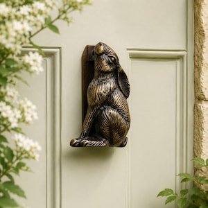May include: A bronze-colored door knocker shaped like a seated hare, mounted on a light green door. The hare is detailed with textured fur and is looking upwards. The door has paneling and is framed by white flowers and greenery.