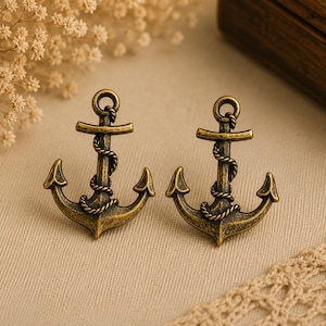 Könnte beinhalten: Zwei bronzefarbene Anker-Ornamente. Jeder Anker hat ein Seildetail und einen Ring oben. Die Anker werden auf einer beigen Oberfläche mit Trockenblumen und einer Holzkiste im Hintergrund präsentiert.