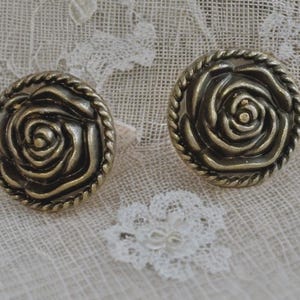 May include: A pair of antique brass-coloured buttons, each with a rose design and a rope-style border. The buttons are set on a textured, off-white fabric with delicate lace details. The buttons are approximately 3 cm in diameter.