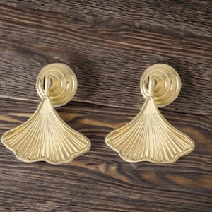 May include: Two gold cabinet pulls with a fan-shaped design. Each pull has a circular backplate and a detailed, textured fan shape. The pulls are made of metal and are set against a dark wood grain background.