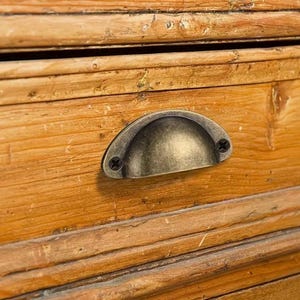 May include: Close-up of a wooden drawer with a warm, weathered finish. It features a semi-circular, antique-style metal handle, secured with two screws. The wood grain is visible, adding to the rustic aesthetic.