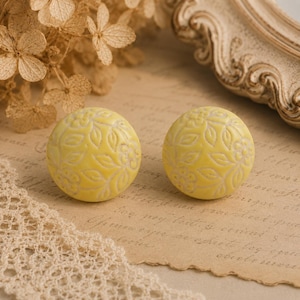 Può includere: Due pomelli decorativi rotondi gialli con un motivo floreale intagliato. I pomelli sono posizionati su uno sfondo in stile vintage con fiori secchi e una cornice decorativa.