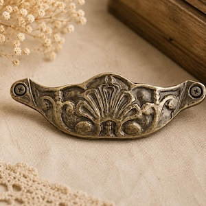 May include: Antique brass drawer pull with an ornate, symmetrical design featuring a central fan-like motif. The pull is secured with two screws and has a weathered finish, suggesting vintage charm. The hardware is likely for a cabinet or drawer.