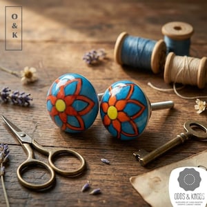 May include: Two blue ceramic cabinet knobs with a red and yellow floral design. The knobs are set on a wooden surface with sewing supplies, a key, and the text "Odds & Knobs".