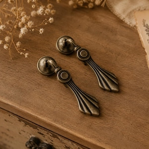 Può includere: Due pomelli per cassetti antichi in bronzo. Ogni pomello presenta un pomolo rotondo e una maniglia decorativa a forma di ventaglio. I pomelli sono appoggiati su una superficie di legno, suggerendo un'estetica vintage o rustica.